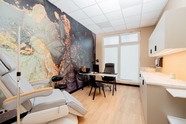 Gabinet FreeMindClinic z szarym fotelem do przeprowadzania leczenia Ketaminą. Po lewej fotel i kolorowa ściana. Po prawej szafki, na końcu biurko oraz duże okno.